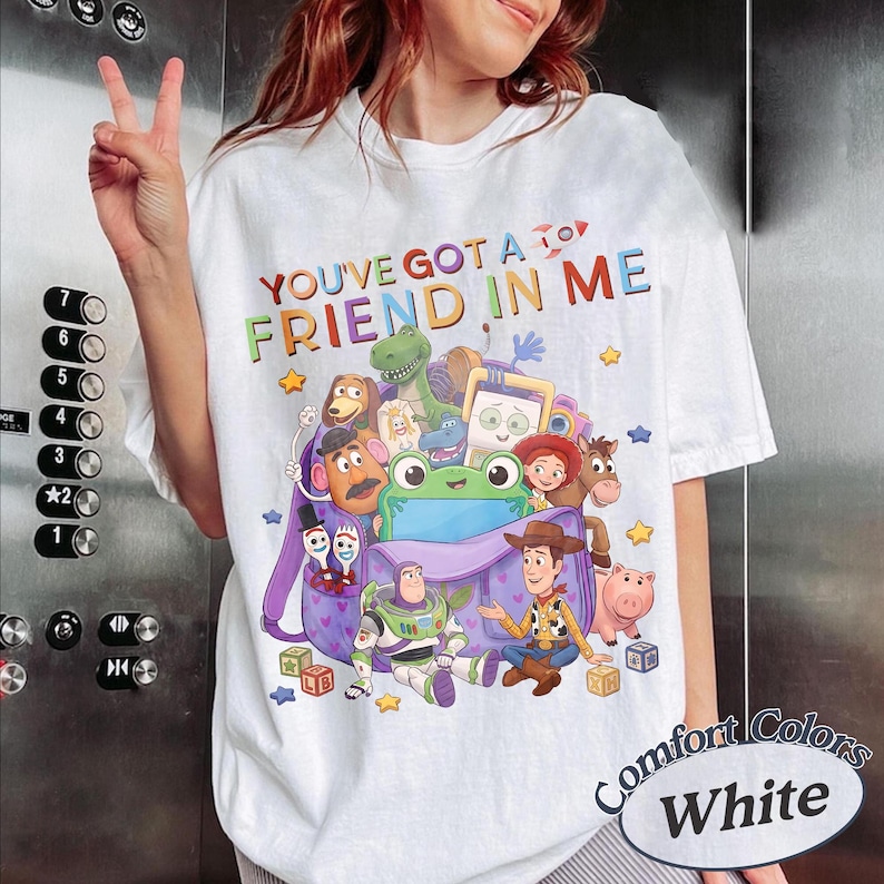 May include: A white t-shirt featuring a colorful graphic of various animated characters from the Toy Story franchise emerging from a purple backpack, with the text "YOU'VE GOT A FRIEND IN ME" printed in multicolored letters above the illustration.