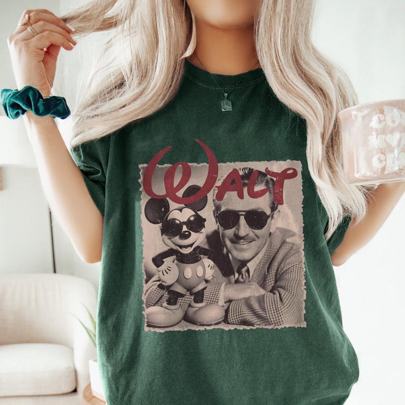 May include: Dark green t-shirt featuring a vintage-style graphic of Mickey Mouse wearing sunglasses next to Walt Disney, with the name "WALT" in red. The shirt is a crew neck and has short sleeves.