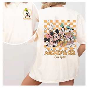 May include: A white t-shirt with a checkered pattern and a graphic of Mickey Mouse and his friends. The graphic includes Mickey Mouse, Minnie Mouse, Donald Duck, Goofy, Pluto, and Daisy Duck. The text "MICKEY & CO. Est. 1928" is printed below the graphic.