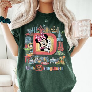 May include: Dark green t-shirt featuring a colorful graphic of Minnie Mouse with a pink polka dot bow, set within a retro-style frame. The shirt also displays the text "Walt Disney World" in a rainbow font, surrounded by iconic park imagery.