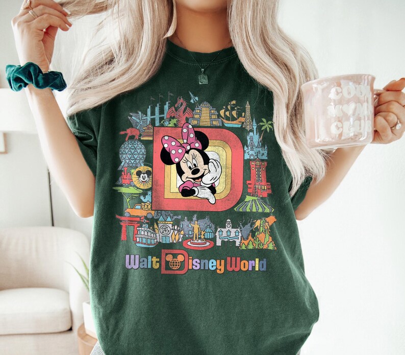 May include: Dark green t-shirt featuring a colorful graphic of Minnie Mouse with a pink polka dot bow, set within a retro-style frame. The shirt also displays the text "Walt Disney World" in a rainbow font, surrounded by iconic park imagery.