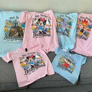 May include: Collection of light blue and pink t-shirts featuring various Disney characters and the Disneyland logo. Designs include Minnie Mouse, The Mandalorian, and other park-themed graphics. The shirts are displayed on a gray couch.
