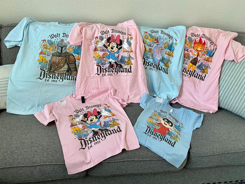 May include: Collection of light blue and pink t-shirts featuring various Disney characters and the Disneyland logo. Designs include Minnie Mouse, The Mandalorian, and other park-themed graphics. The shirts are displayed on a gray couch.