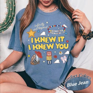 May include: A blue jean coloured t-shirt featuring a graphic print with the text "infinity and beyond", "YOU'VE GOT A FRIEND IN ME", "I KNEW IT", and "I KNEW YOU". The design includes a yellow star, a beaded bracelet, a cowboy boot, and alphabet blocks.