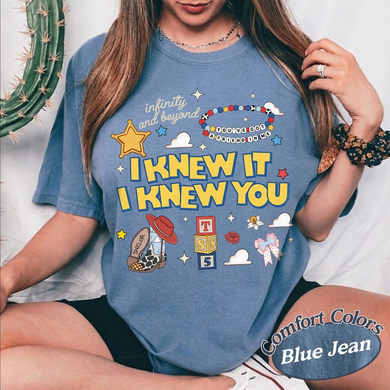May include: A blue jean coloured t-shirt featuring a graphic print with the text "infinity and beyond", "YOU'VE GOT A FRIEND IN ME", "I KNEW IT", and "I KNEW YOU". The design includes a yellow star, a beaded bracelet, a cowboy boot, and alphabet blocks.