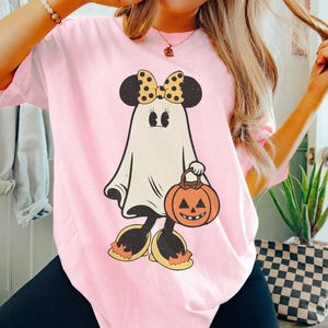May include: Pink t-shirt with a cartoon ghost wearing a yellow and black bow and carrying a pumpkin. The ghost has large eyes and a friendly smile.