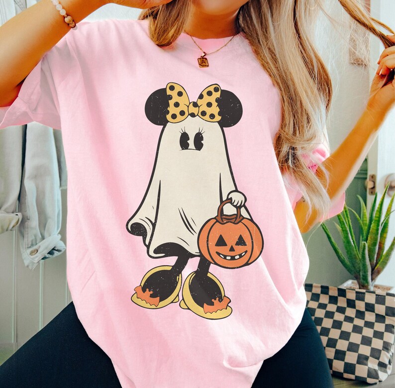 May include: Pink t-shirt with a cartoon ghost wearing a yellow and black bow and carrying a pumpkin. The ghost has large eyes and a friendly smile.