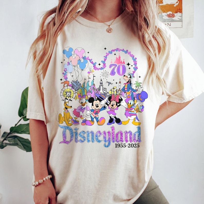May include: Cream-colored t-shirt featuring a colorful graphic celebrating Disneyland's 70th anniversary. The design includes Mickey and Minnie Mouse, Donald Duck, Goofy, and other characters, along with the text "Disneyland 1955-2025".