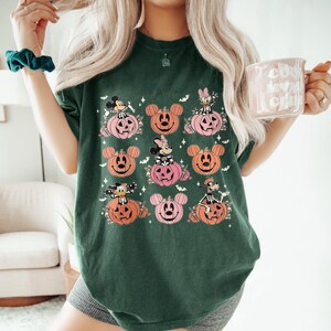 May include: Dark green t-shirt featuring a Halloween-themed design. The design includes cartoon characters on pink and orange pumpkin faces. The shirt is a comfortable, casual style, perfect for the fall season.