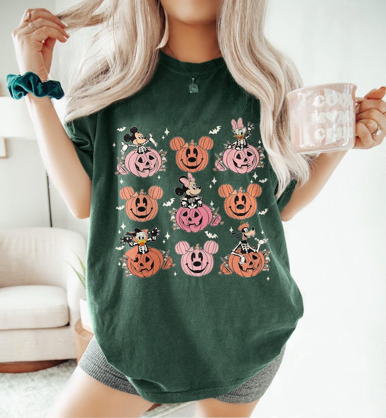 May include: Dark green t-shirt featuring a Halloween-themed design. The design includes cartoon characters on pink and orange pumpkin faces. The shirt is a comfortable, casual style, perfect for the fall season.