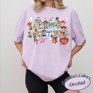 May include: A light purple short-sleeve t-shirt featuring a graphic print of Toy Story characters including Woody, Buzz Lightyear, Jessie, Rex, Bullseye, and Hamm. The text reads I KNEW IT I KNEW YOU with a heart shape containing the letters TS.