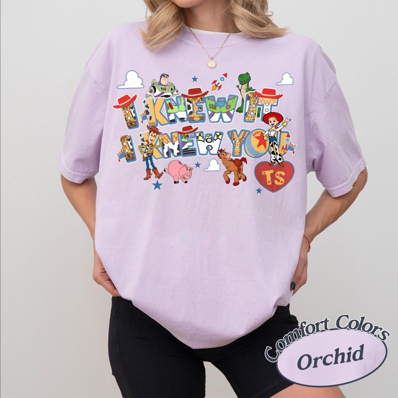 May include: A light purple short-sleeve t-shirt featuring a graphic print of Toy Story characters including Woody, Buzz Lightyear, Jessie, Rex, Bullseye, and Hamm. The text reads I KNEW IT I KNEW YOU with a heart shape containing the letters TS.