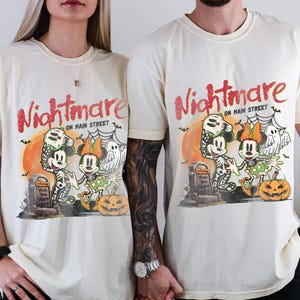 May include: Ivory-colored t-shirts featuring a Halloween-themed graphic with Mickey and Minnie Mouse dressed as skeletons. The design includes the text "Nightmare on Main Street", a pumpkin, and a tombstone. The shirts are short-sleeved.