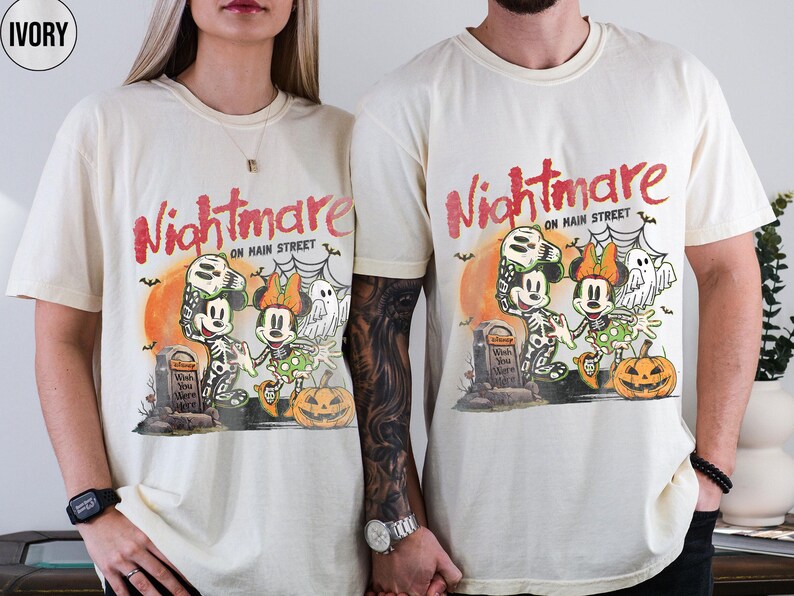 May include: Ivory-colored t-shirts featuring a Halloween-themed graphic with Mickey and Minnie Mouse dressed as skeletons. The design includes the text "Nightmare on Main Street", a pumpkin, and a tombstone. The shirts are short-sleeved.