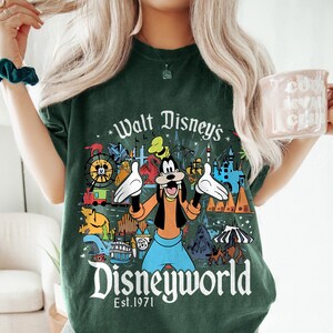 May include: Dark green t-shirt featuring a colorful graphic of Goofy with the text "Walt Disney's Disneyworld Est. 1971". The graphic includes various Disney park icons. The shirt is a comfortable, casual style.