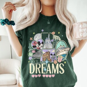May include: Dark green t-shirt with a colorful graphic featuring Mickey and Minnie Mouse, a castle, and a cruise ship. The text "What Dreams Are Made Of" is displayed below the graphic. The shirt has a crew neck and short sleeves.