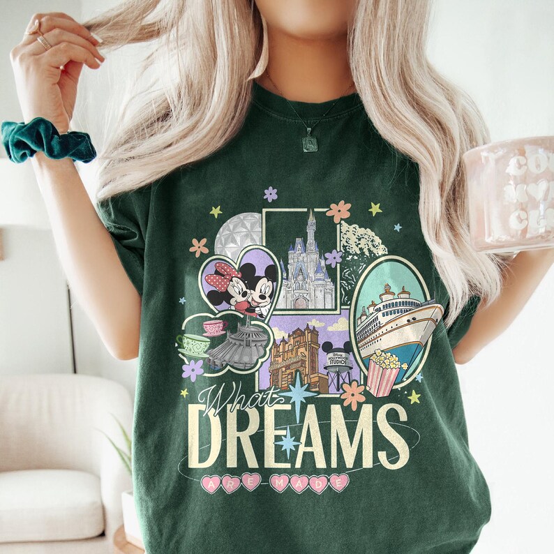 May include: Dark green t-shirt with a colorful graphic featuring Mickey and Minnie Mouse, a castle, and a cruise ship. The text "What Dreams Are Made Of" is displayed below the graphic. The shirt has a crew neck and short sleeves.