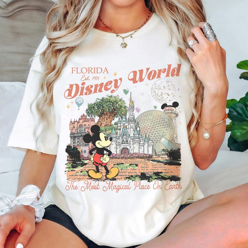 May include: Cream-colored t-shirt featuring a colorful graphic of Disney World landmarks, including Cinderella's castle and Mickey Mouse. The text "Florida Disney World" and "The Most Magical Place On Earth" are printed on the shirt.