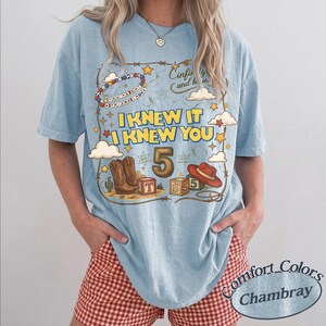 May include: A light blue t-shirt featuring a graphic print with the text I KNEW IT I KNEW YOU, a large number 5, cowboy boots, a red cowboy hat, alphabet blocks, clouds, stars, and a beaded bracelet with the text YOU'VE GOT A FRIEND IN ME.
