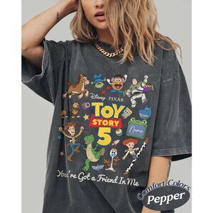 May include: A charcoal gray t-shirt featuring a colorful graphic print of Toy Story characters including Woody, Buzz Lightyear, Jessie, Rex, and Forky. The design includes the text Toy Story 5 and You've Got a Friend In Me, with a blank green sign.