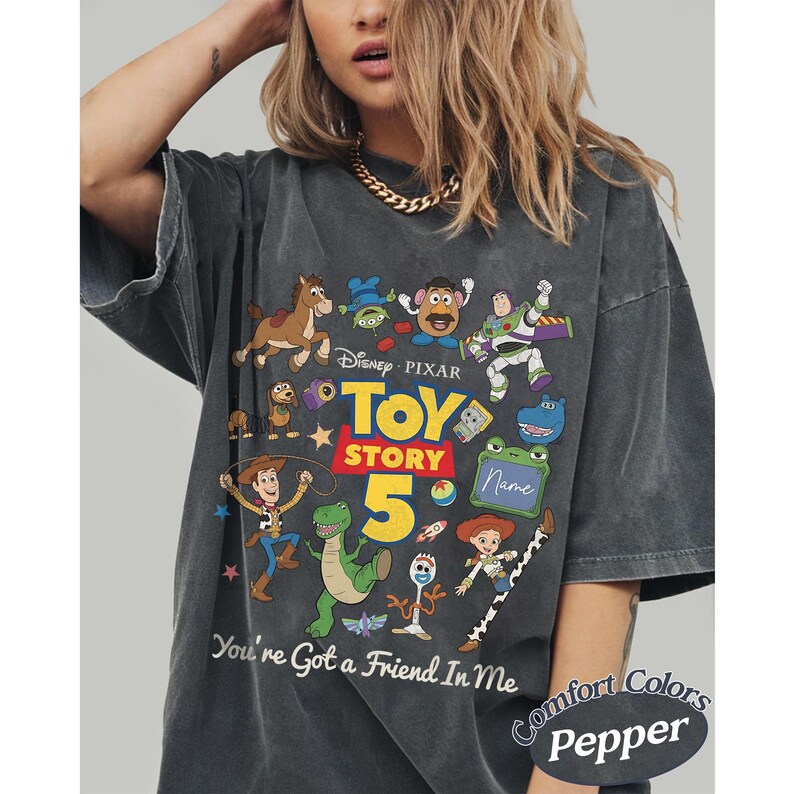 May include: A charcoal gray t-shirt featuring a colorful graphic print of Toy Story characters including Woody, Buzz Lightyear, Jessie, Rex, and Forky. The design includes the text Toy Story 5 and You've Got a Friend In Me, with a blank green sign.