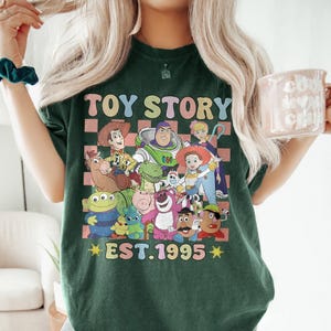 May include: Green t-shirt with a checkered background featuring characters from the Toy Story franchise. The text "TOY STORY EST. 1995" is printed on the shirt.
