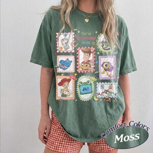 May include: A moss green short-sleeve t-shirt featuring a graphic print of various cartoon characters arranged in postage stamp frames. The text "You've Got A Friend In Me" is printed in the center. The shirt has a crew neckline and a loose fit.