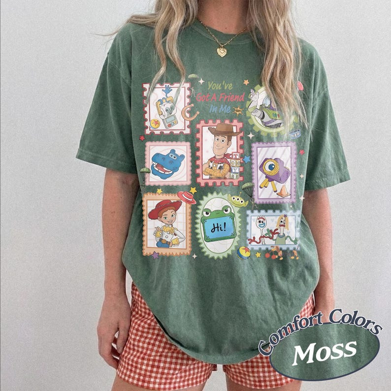 May include: A moss green short-sleeve t-shirt featuring a graphic print of various cartoon characters arranged in postage stamp frames. The text "You've Got A Friend In Me" is printed in the center. The shirt has a crew neckline and a loose fit.