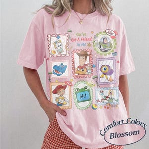 May include: A light pink t-shirt featuring a graphic print with the text "You've Got A Friend In Me" surrounded by various postage stamp-style frames containing illustrations of characters and items from the Toy Story film franchise.