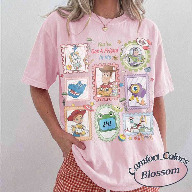 May include: A light pink t-shirt featuring a graphic print with the text "You've Got A Friend In Me" surrounded by various postage stamp-style frames containing illustrations of characters and items from the Toy Story film franchise.