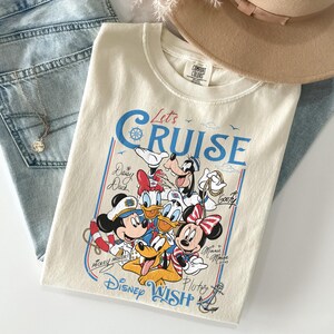 May include: Cream-colored t-shirt featuring a graphic of Disney characters, including Mickey Mouse, Minnie Mouse, Donald Duck, and Goofy. The shirt has the text "Let's Cruise" in blue and red, with the words "Disney Wish" at the bottom.