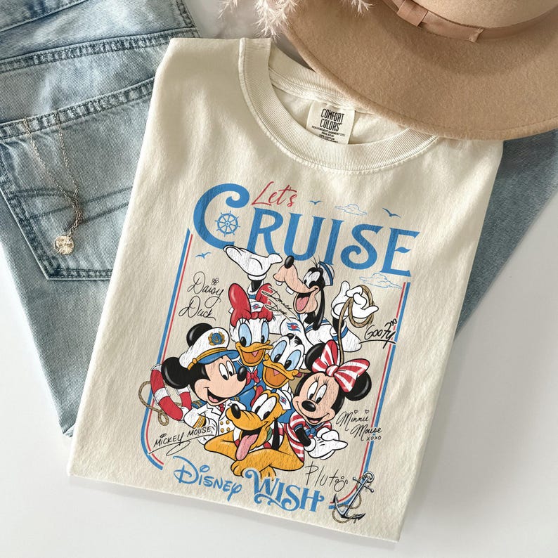 May include: Cream-colored t-shirt featuring a graphic of Disney characters, including Mickey Mouse, Minnie Mouse, Donald Duck, and Goofy. The shirt has the text "Let's Cruise" in blue and red, with the words "Disney Wish" at the bottom.