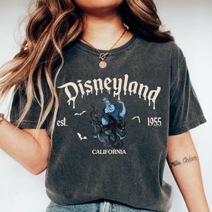 K&ouml;nnte beinhalten: Dunkelgraues T-Shirt mit dem Wort "Disneyland" in tropfender wei&szlig;er Schrift. Das Shirt zeigt eine blau-schwarze Grafik einer Figur, den Text "est. 1955 CALIFORNIA" und mehrere Fledermaus-Silhouetten.