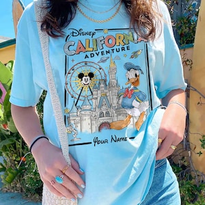 Puede incluir: Camiseta azul claro con un gr&aacute;fico de Disney California Adventure. El dise&ntilde;o incluye a Mickey Mouse en una noria, el Pato Donald y un castillo. El texto "California Adventure" se muestra en una fuente estilizada.