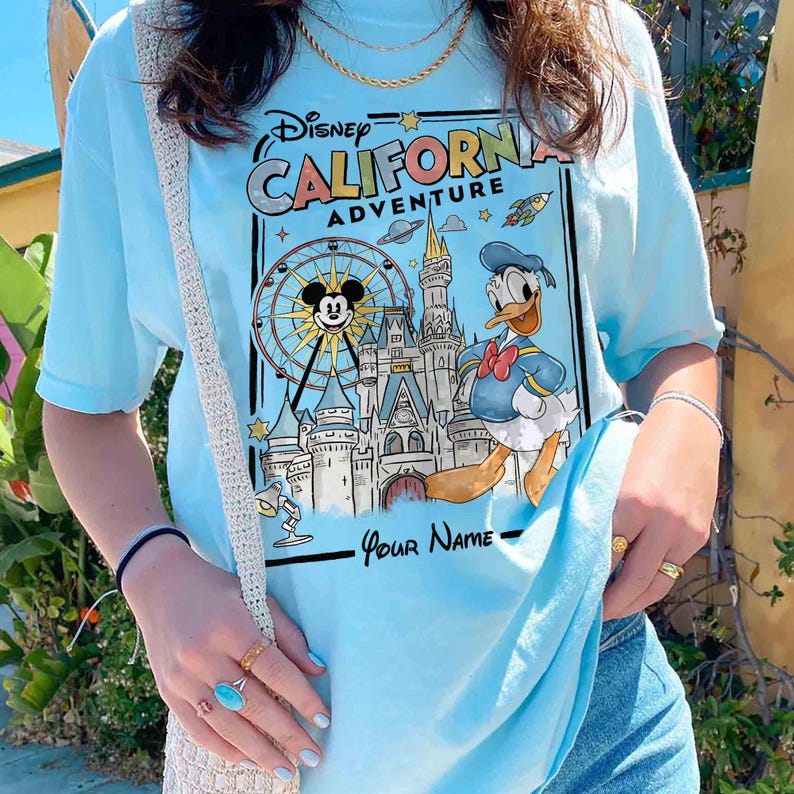 Puede incluir: Camiseta azul claro con un gr&aacute;fico de Disney California Adventure. El dise&ntilde;o incluye a Mickey Mouse en una noria, el Pato Donald y un castillo. El texto "California Adventure" se muestra en una fuente estilizada.