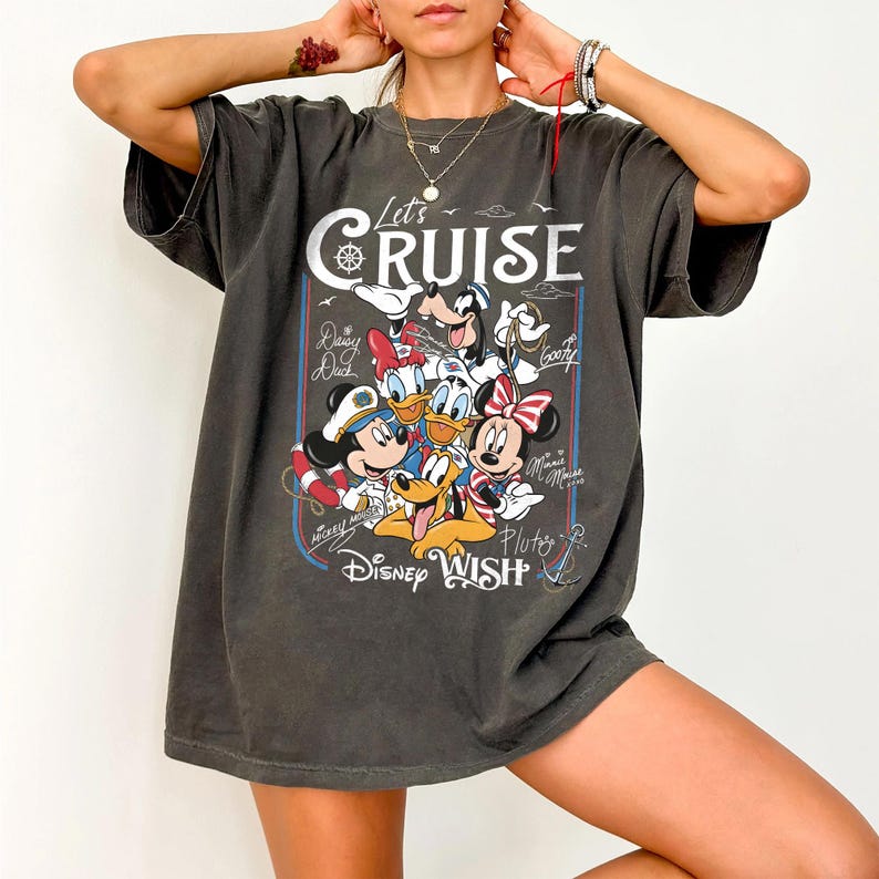May include: Dark gray t-shirt featuring a graphic of Disney characters, including Mickey Mouse, Minnie Mouse, Donald Duck, and Goofy. The shirt has the text "Let's Cruise" and "Disney Wish".