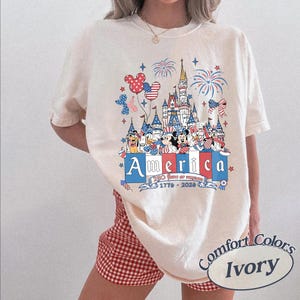 May include: An ivory t-shirt featuring a graphic print of a castle, fireworks, and cartoon characters in red, white, and blue. The text reads America, 250 Years of Freedom, and 1776-2026. The shirt is paired with red and white gingham shorts.