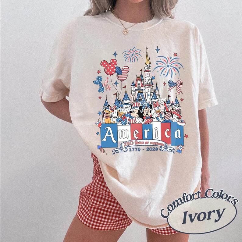 May include: An ivory t-shirt featuring a graphic print of a castle, fireworks, and cartoon characters in red, white, and blue. The text reads America, 250 Years of Freedom, and 1776-2026. The shirt is paired with red and white gingham shorts.