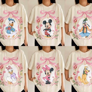 May include: Cream-colored t-shirts featuring Disney characters: Donald Duck, Mickey Mouse, Goofy, Daisy Duck, Minnie Mouse, and Pluto. Each character is framed in an oval with a pink bow and floral accents. The shirts have short sleeves.