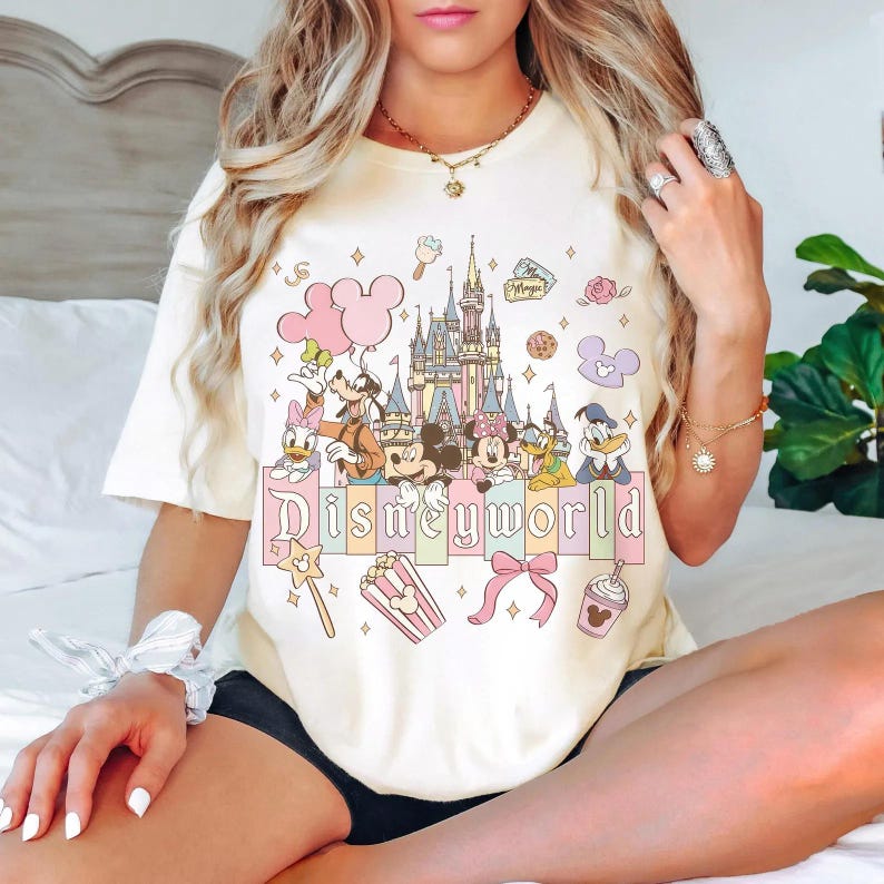 May include: Cream-colored t-shirt featuring a colorful Disneyworld graphic. The design includes Disney characters, a castle, and the word "Disneyworld" in a pastel font. Other elements include balloons, popcorn, and a magic wand.