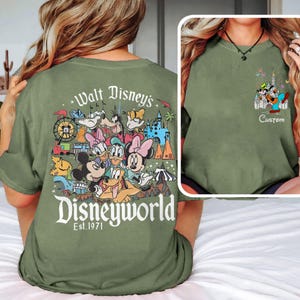 May include: Olive green t-shirt featuring a colorful Disney World graphic on the back with Mickey Mouse, Donald Duck, and other characters. The front has a smaller graphic with Goofy, Mickey, and a castle. Text reads "Custom".