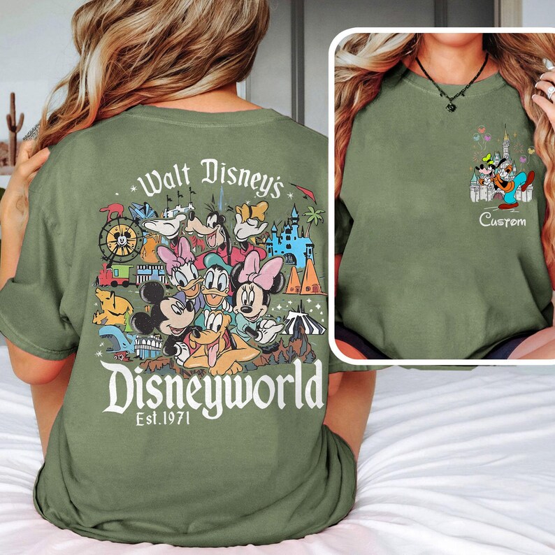 May include: Olive green t-shirt featuring a colorful Disney World graphic on the back with Mickey Mouse, Donald Duck, and other characters. The front has a smaller graphic with Goofy, Mickey, and a castle. Text reads "Custom".