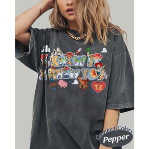 May include: A charcoal gray short-sleeve t-shirt featuring a graphic print with the text "I KNEW IT I KNEW YOU" and various cartoon characters. The design includes colorful illustrations of a cowboy, space ranger, pig, horse, and dinosaur.