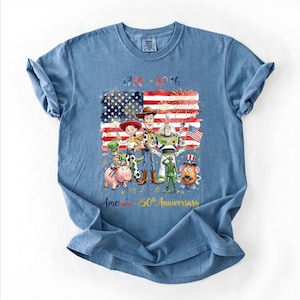 May include: A blue t-shirt featuring a graphic of Toy Story characters including Woody, Buzz Lightyear, Jessie, Rex, Hamm, Mr. Potato Head, and a green soldier in front of an American flag with fireworks. Text reads 1776-2026 America 250th Anniversary.