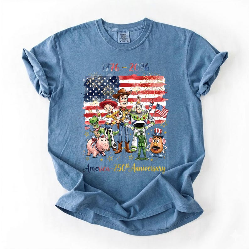 May include: A blue t-shirt featuring a graphic of Toy Story characters including Woody, Buzz Lightyear, Jessie, Rex, Hamm, Mr. Potato Head, and a green soldier in front of an American flag with fireworks. Text reads 1776-2026 America 250th Anniversary.