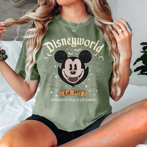 May include: Olive green t-shirt with a distressed "Disneyworld" graphic featuring Mickey Mouse with fangs. The design includes the text "Est. 1971" and "Spookiest Place on Earth" on a banner.