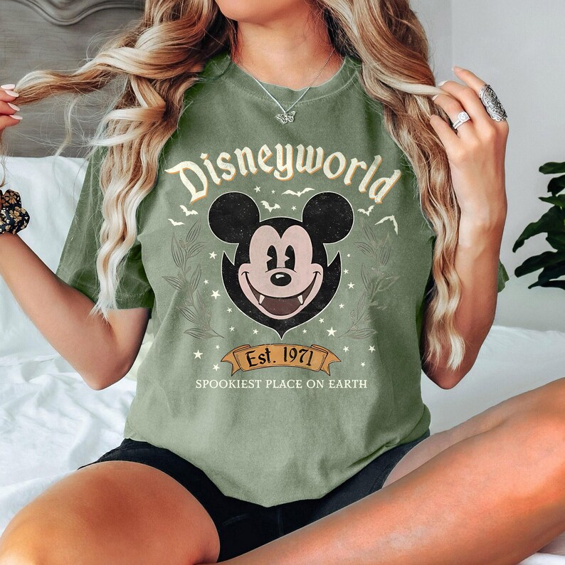 May include: Olive green t-shirt with a distressed "Disneyworld" graphic featuring Mickey Mouse with fangs. The design includes the text "Est. 1971" and "Spookiest Place on Earth" on a banner.
