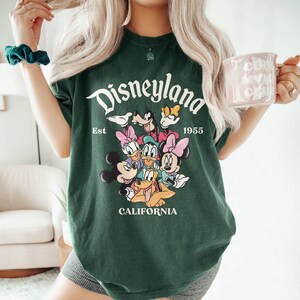 May include: Dark green t-shirt with a Disneyland graphic featuring Mickey Mouse, Minnie Mouse, Donald Duck, Daisy Duck, Goofy, and Pluto. The text "Disneyland Est 1955 California" is printed in white. The shirt is a casual style.