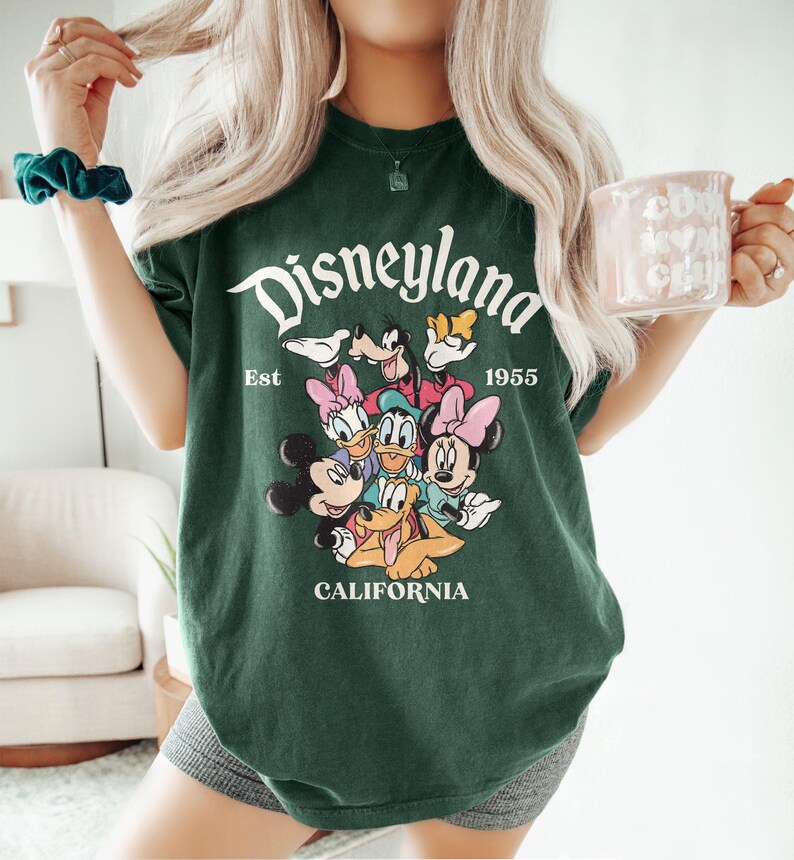 May include: Dark green t-shirt with a Disneyland graphic featuring Mickey Mouse, Minnie Mouse, Donald Duck, Daisy Duck, Goofy, and Pluto. The text "Disneyland Est 1955 California" is printed in white. The shirt is a casual style.