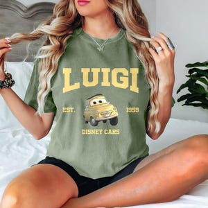 Puede incluir: Camiseta verde de manga corta con un coche de dibujos animados amarillo y el texto "LUIGI EST. 1959 DISNEY CARS".